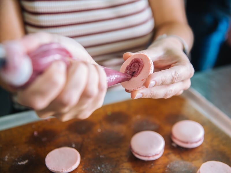 Person piping a macaron shell with pink filling